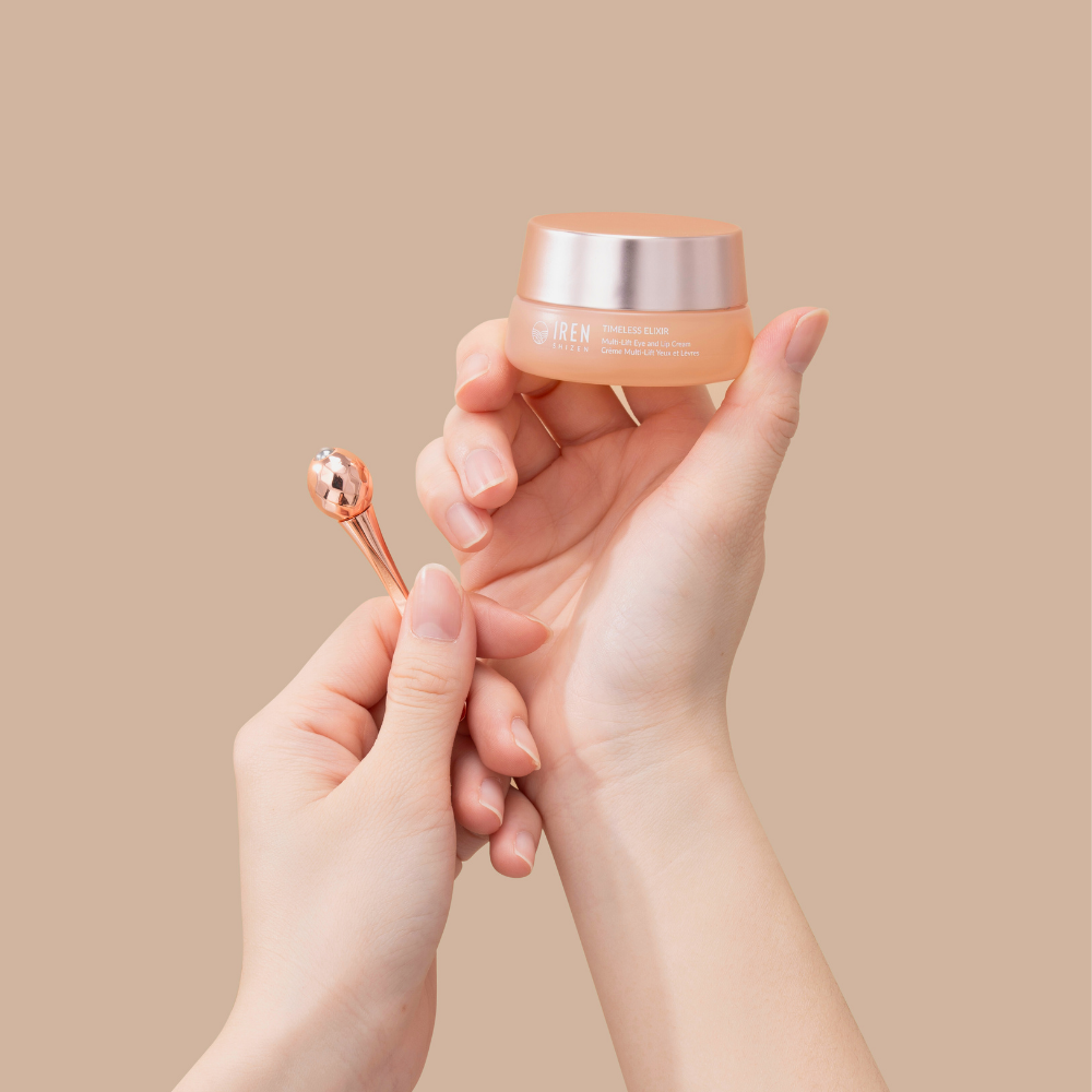 Hands holding a small jar of IREN Shizen TIMELESS ELIXIR Multi-Lift Eye and Lip Cream and a metal applicator against a beige background, perfect for eye area rejuvenation.