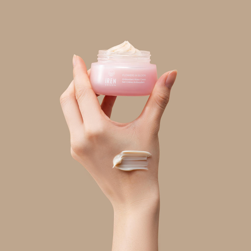A hand holding a jar of FLOWERS IN BLOOM Antioxidant Water Cream by IREN Shizen, enriched with natural origin ingredients, with some cream on the fingers against a neutral background.