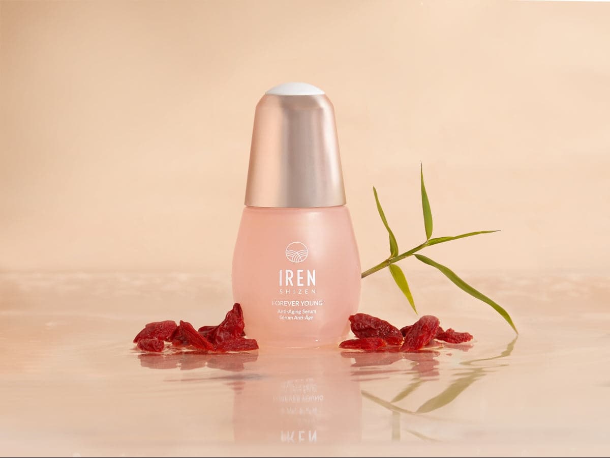 Close-up of the IREN Shizen APAC TIMELESS ELIXIR Anti-Aging Eye Skincare Kit bottle surrounded by dried red berries and a green leaf on a reflective surface with a beige background, showcasing the allure of anti-aging skincare.