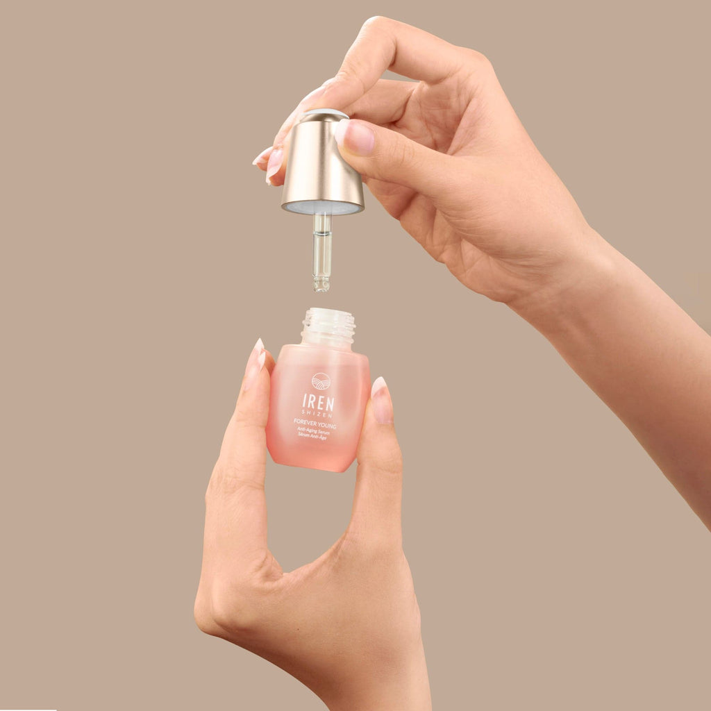 A hand is holding a bottle of GLOW UP Anti-Aging Set, a custom skincare product from IREN Shizen, on a beige background.