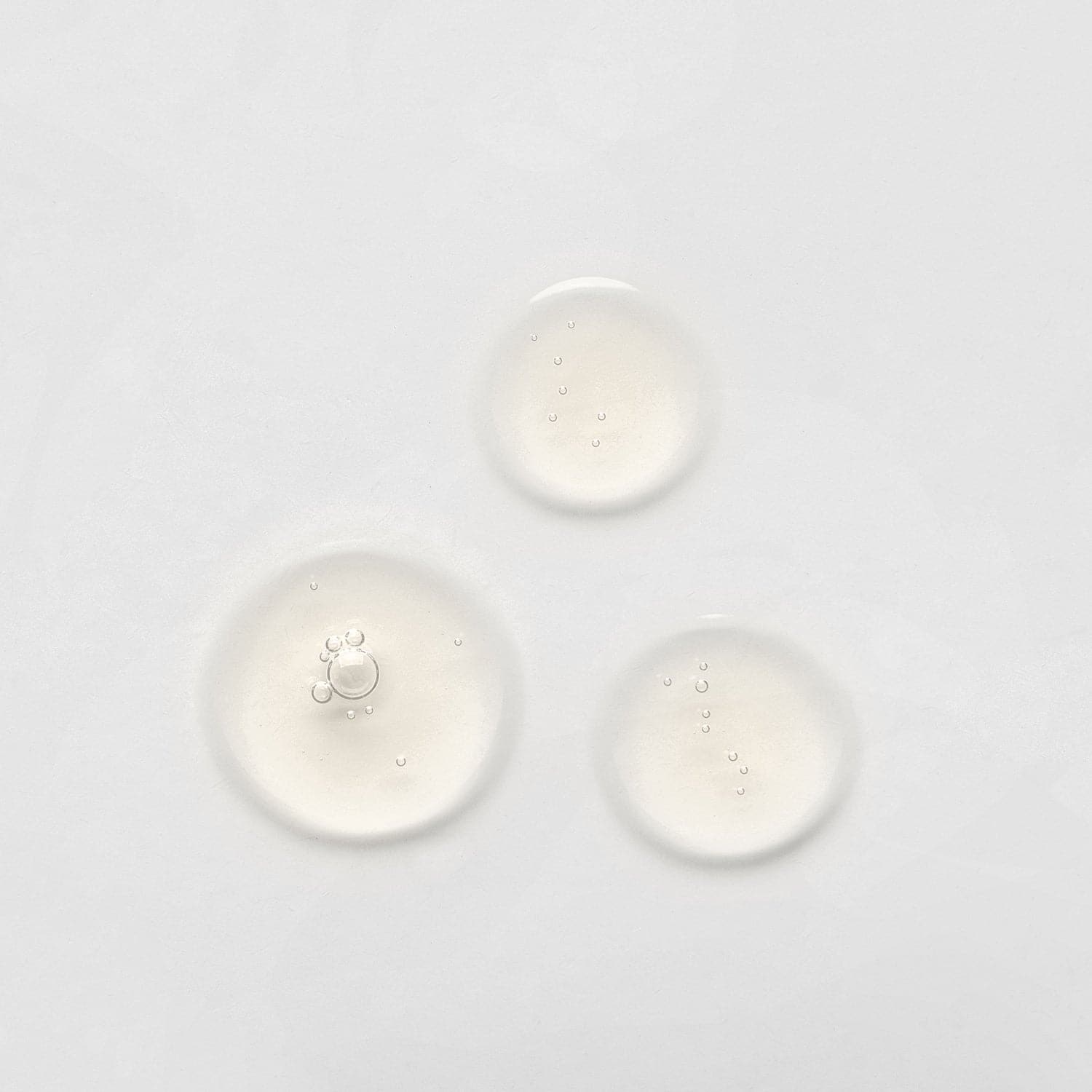 Three small, clear liquid droplets on a plain white surface, possibly from the LIFT & FIRM Anti-Aging Duo Serum Set by IREN Shizen, which contains Argireline Peptide.