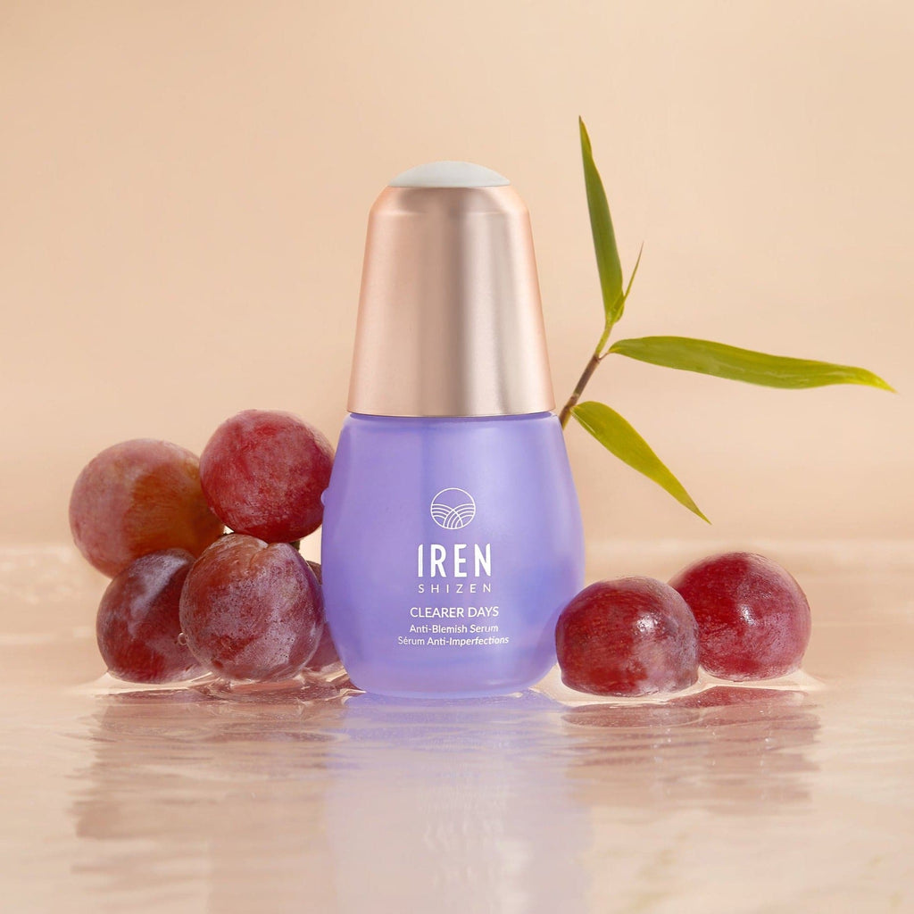 A bottle of CUSTOMIZED skincare, CLEARER DAYS Anti-Blemish Serum by IREN Shizen, with grapes and onsen skincare next to it.